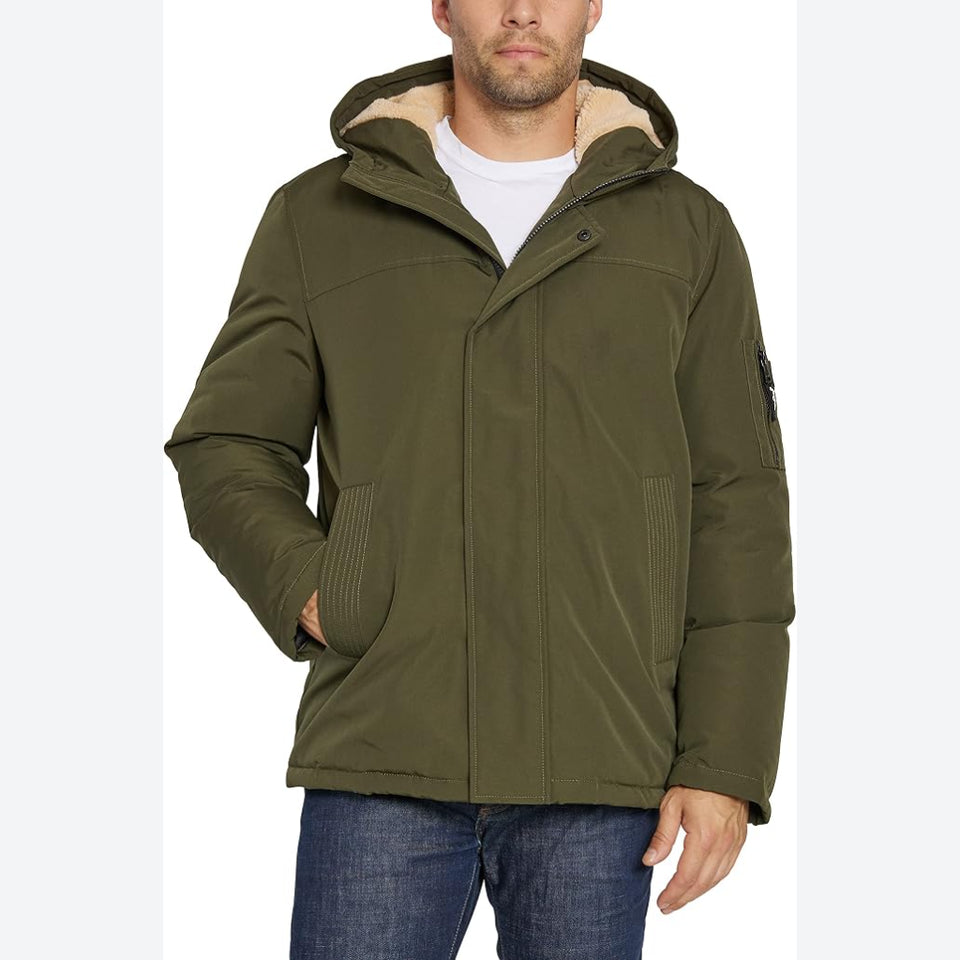A man wearing an olive green hooded winter jacket with a beige sherpa-lined hood, paired with a white t-shirt and blue jeans, shown against a plain white background.