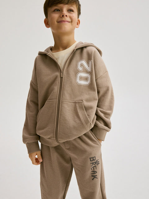 Child wearing a brown tracksuit with '20' and 'BREAK' text on a white background