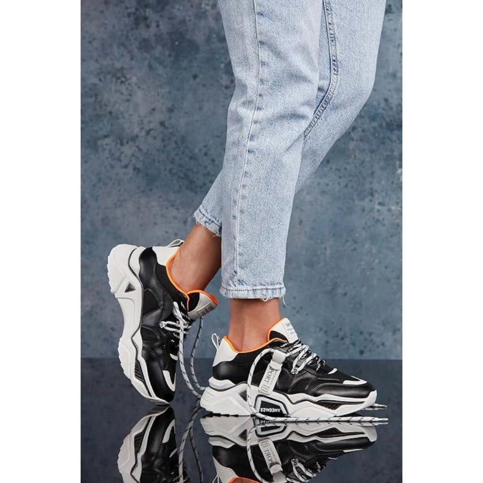 A person wearing black and white chunky platform sneakers with bright orange lining, paired with light-wash blue jeans, standing on a reflective dark surface against a textured blue-gray background.
