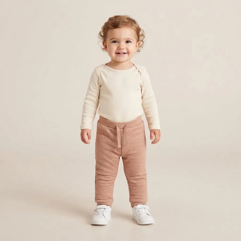 A toddler standing against a warm beige studio background, wearing a cream long-sleeve top, dusty rose drawstring pants, and white sneakers.