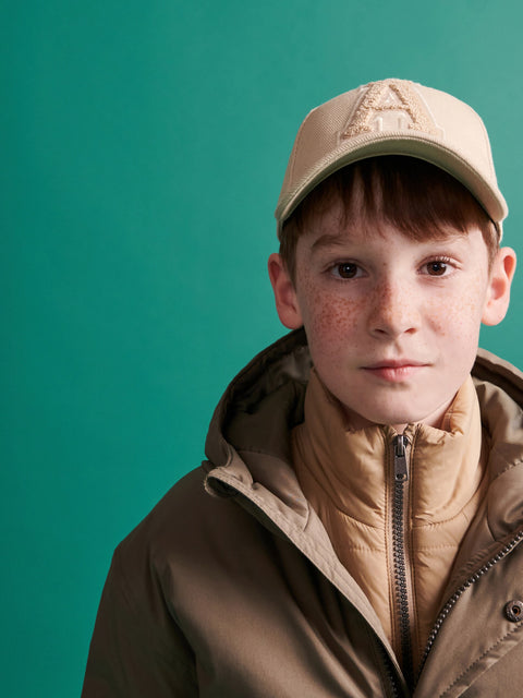Young boy wearing a beige cap and brown jacket against a teal background