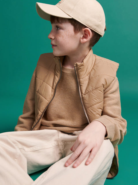 Child wearing a beige quilted jacket and cap against a teal background