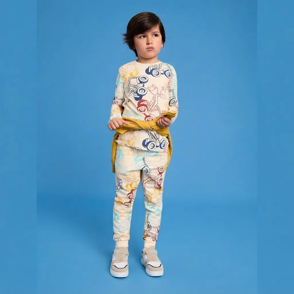 A young boy modeling a matching two-piece pajama set with a colorful abstract print on a cream base, featuring a long-sleeve top and pants, styled with a yellow tie belt and white sneakers against a solid blue background.