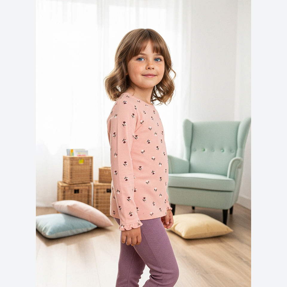 A young girl with wavy brown hair stands in a bright room wearing a light pink long-sleeve top with a small black and white pattern, paired with mauve-purple pants. She is photographed from the side/back, with a mint green armchair, woven storage baskets, and pillows in the background.