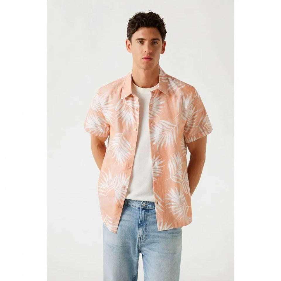 A man wearing a peach short-sleeve button-up shirt with a white palm leaf pattern, layered over a white t-shirt and light blue jeans, standing against a light gray studio background.