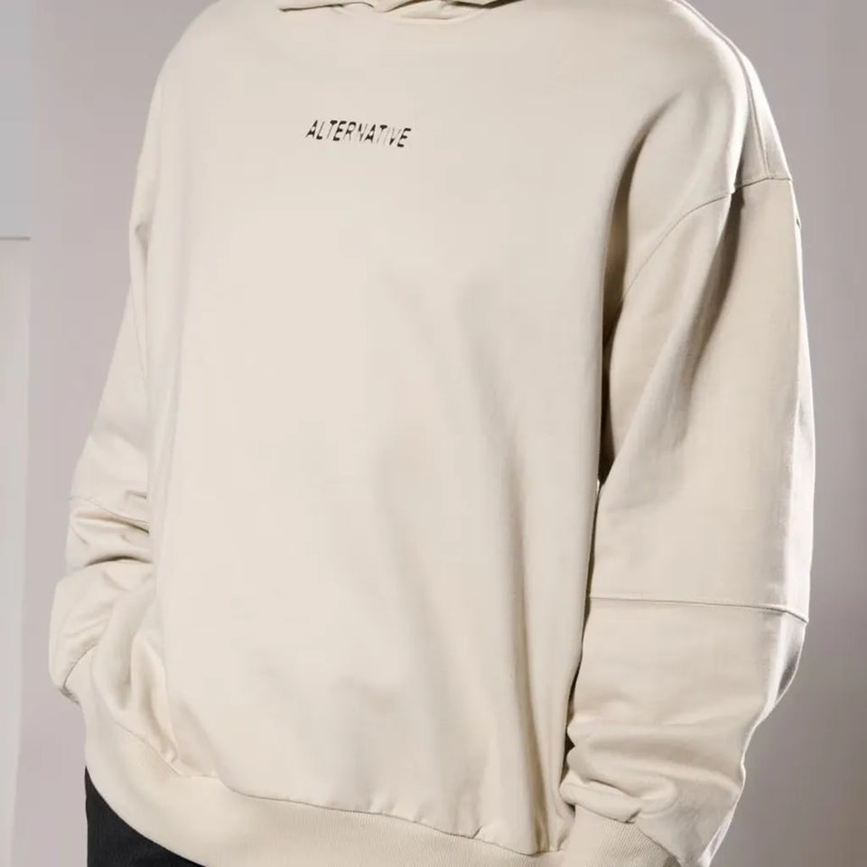 An oversized cream-colored hoodie with 'ALTERNATIVE' printed in black text on the chest, shown on a model against a neutral gray background.