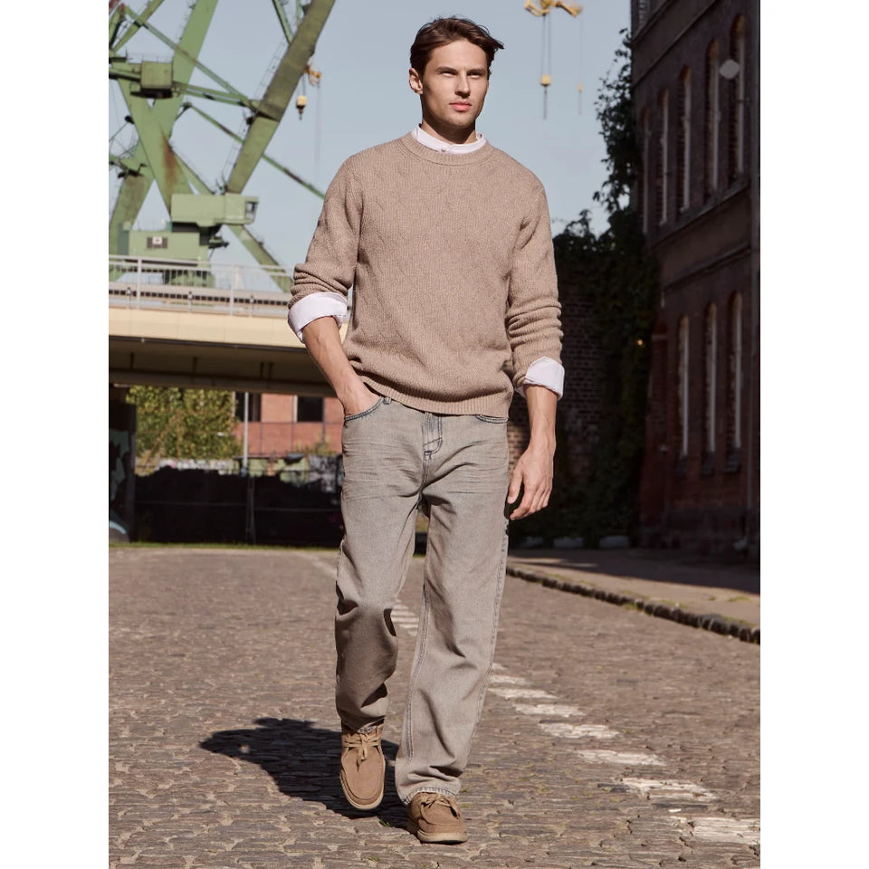 A man walking outdoors on a cobblestone street wearing a beige crewneck sweater over a white collared shirt, beige jeans, and brown shoes.