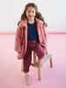 Young girl wearing a pink coat sitting on a small wooden stool against a pink background