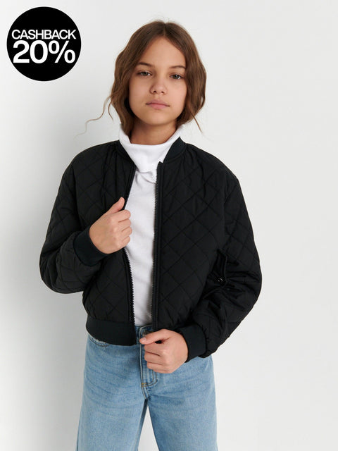 Person wearing a black quilted jacket over a white shirt with light blue jeans on a plain background