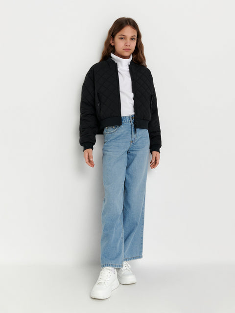 Person wearing a black jacket, white turtleneck, blue jeans, and white sneakers on a white background