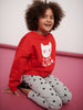 Child wearing a red sweatshirt with a white cat design and text, sitting on a pink surface.