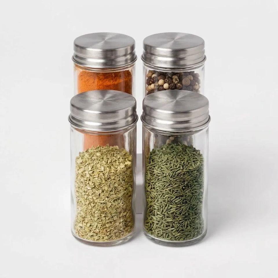 Four clear glass spice jars with silver metal screw-top lids, filled with various whole and ground spices, arranged in a 2x2 grid on a light gray background.