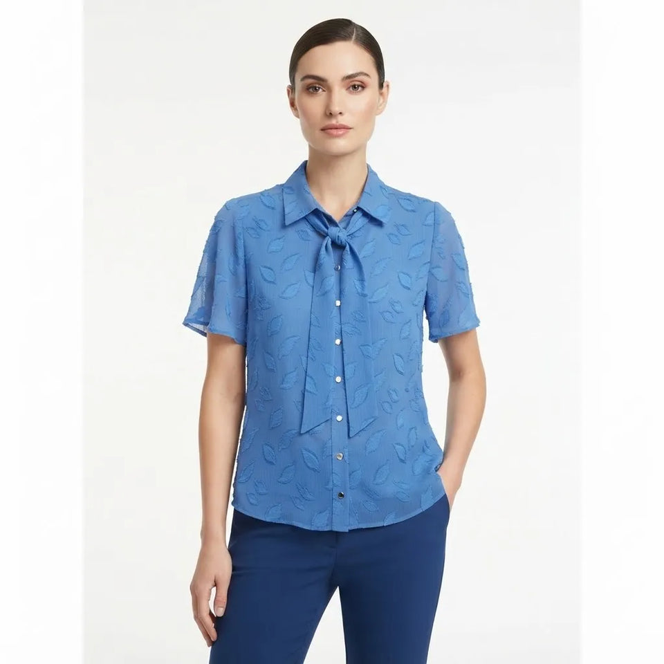 A woman modeling a blue short-sleeve button-up blouse with a tie-neck detail and subtle tonal leaf pattern, paired with dark blue pants, against a plain light background.