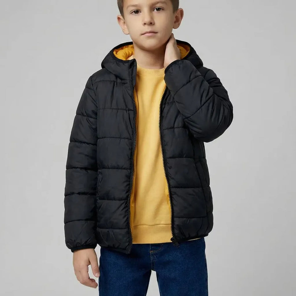 A young boy wearing a black hooded puffer jacket with a yellow lining, layered over a mustard yellow sweatshirt and blue jeans, photographed against a neutral gray studio background.