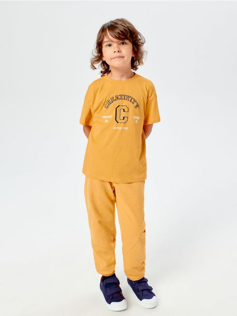 A boy standing against a white background, wearing a mustard yellow t-shirt and matching sweatpants.