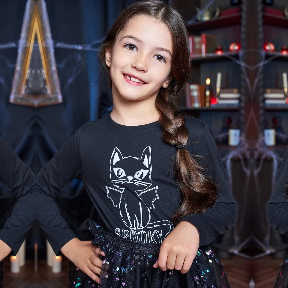 A young girl with a brown braid smiles while wearing a black long-sleeve t-shirt with a white cat graphic and the word 'MOMMY' below it, paired with a sparkly black skirt in a themed indoor setting.