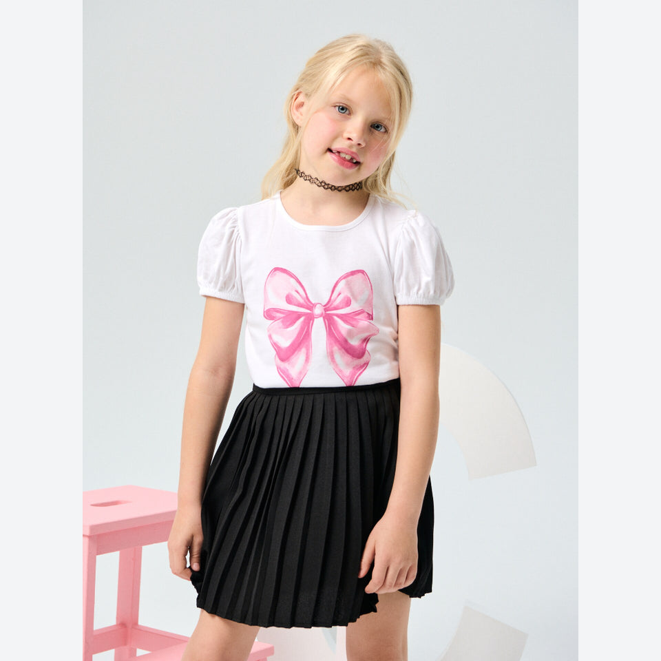 A young girl with blonde hair is wearing a white short-sleeve t-shirt with a pink bow graphic and a black pleated skirt, standing against a light gray background.