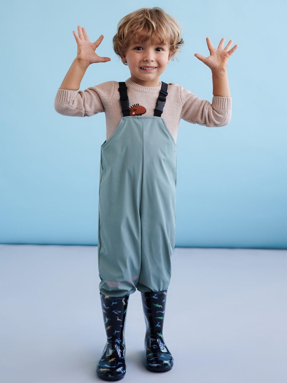 Child wearing a light blue dress overalls with a beige sweater and dark rain boots against a light blue background