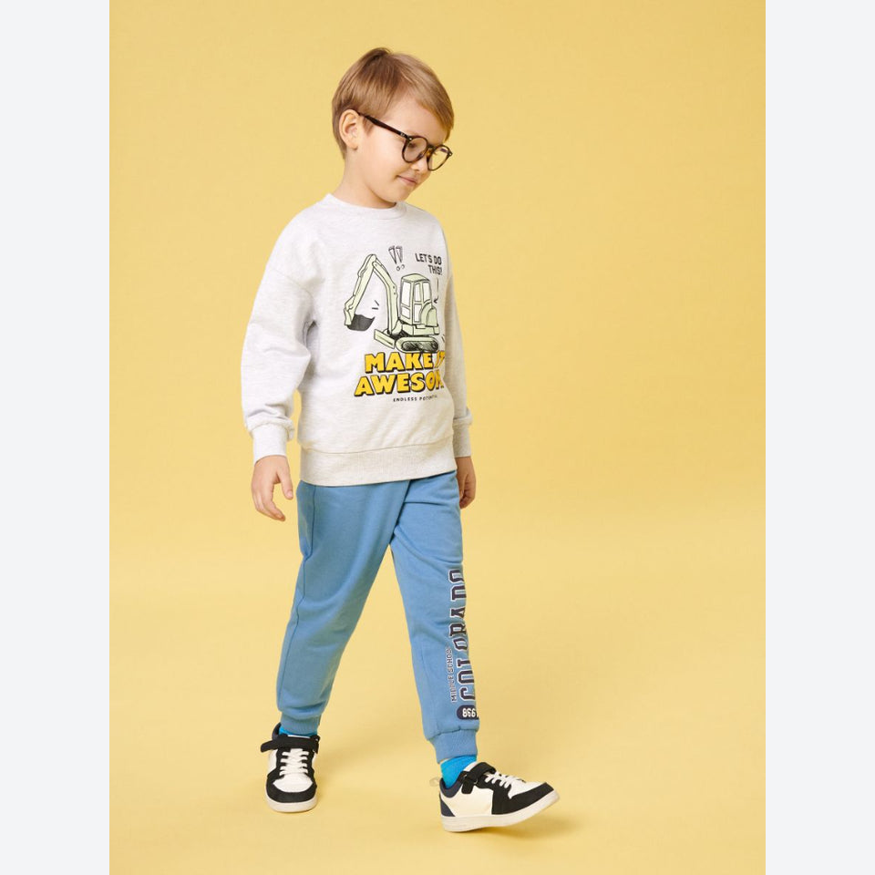 A young boy wearing a white graphic sweatshirt, light blue jogger pants, and black-and-white sneakers, standing against a solid yellow studio background.