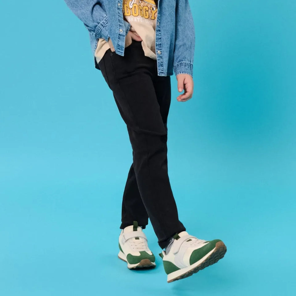A person wearing black slim-fit jeans, a light blue denim jacket, a beige graphic t-shirt, and white sneakers with green accents, walking against a solid bright blue background.
