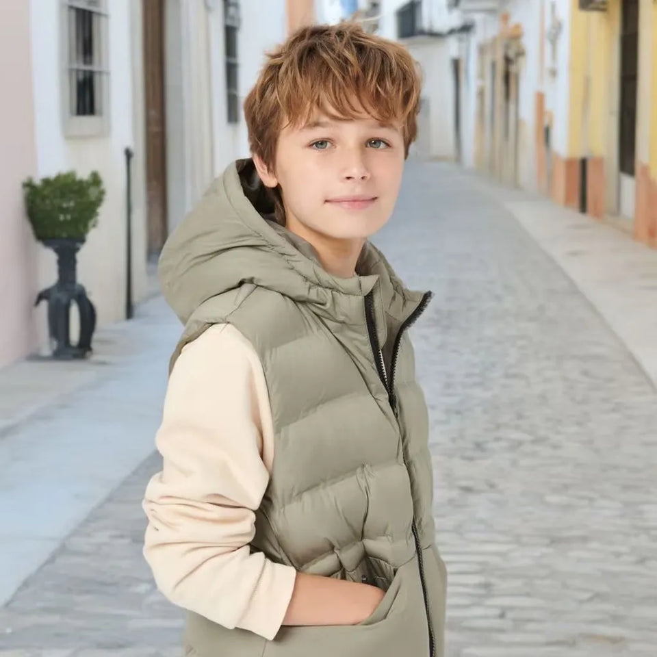 A young boy with light brown hair stands on a cobblestone street wearing a sage green hooded puffer vest over a light beige long-sleeve shirt.