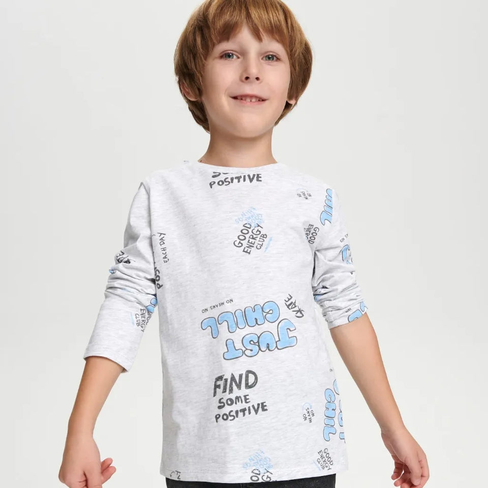 A young boy wearing a light gray heathered long-sleeve t-shirt with blue and black graphic text prints, photographed against a solid light gray background.