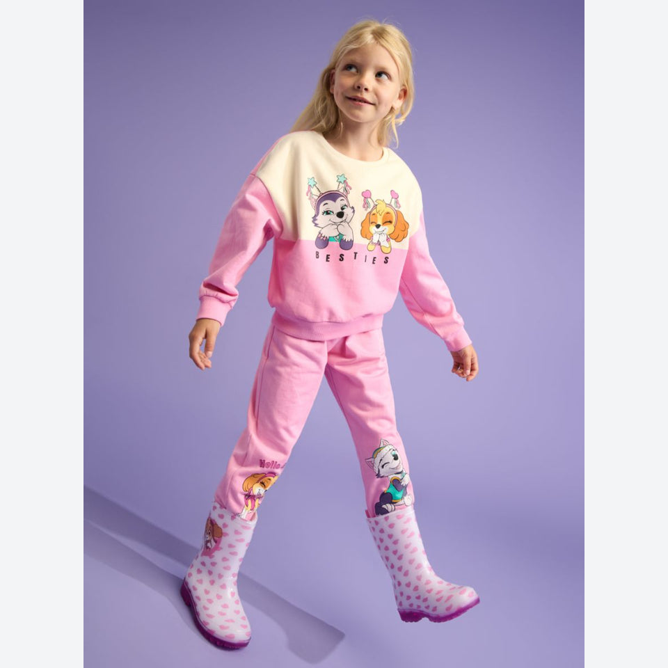 A young girl modeling a pink and cream two-piece tracksuit with cartoon dog graphics, matching pink pants, and pink polka-dot rain boots against a solid lavender studio background.