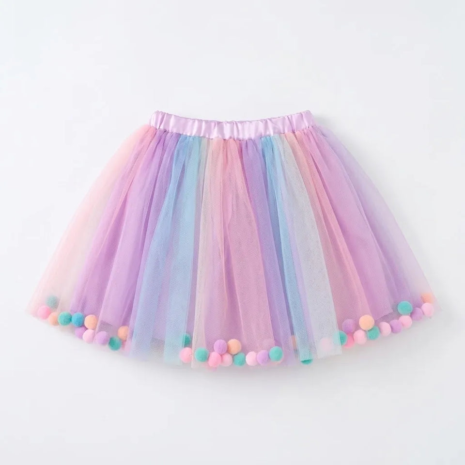 A pastel rainbow tulle tutu skirt with pom-pom trim, featuring vertical layers of pink, purple, blue, and yellow mesh with a matching elastic waistband.