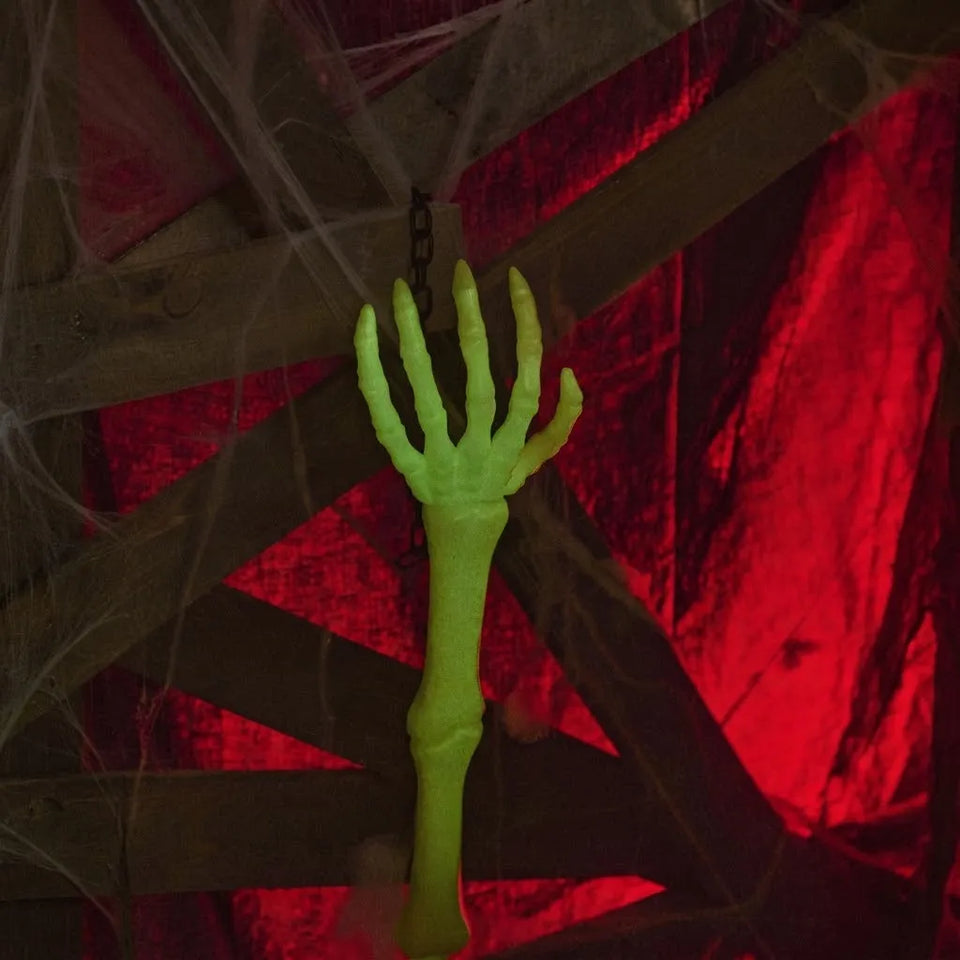 A glowing neon green plastic skeleton hand hanging from a chain, set against a dark background with red lighting and fake cobwebs.