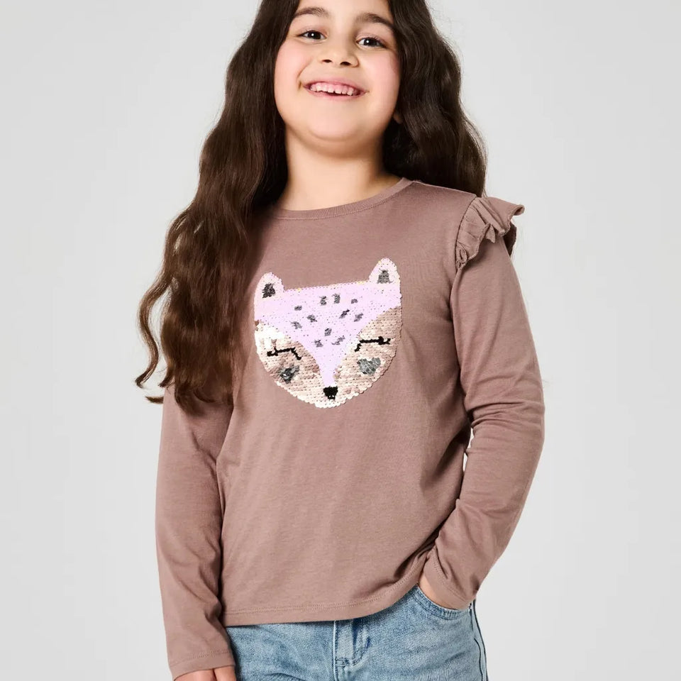 A young girl with long brown hair smiling, wearing a mauve-brown long-sleeve top with a sequined fox face graphic on the front, paired with blue jeans, against a light gray studio background.