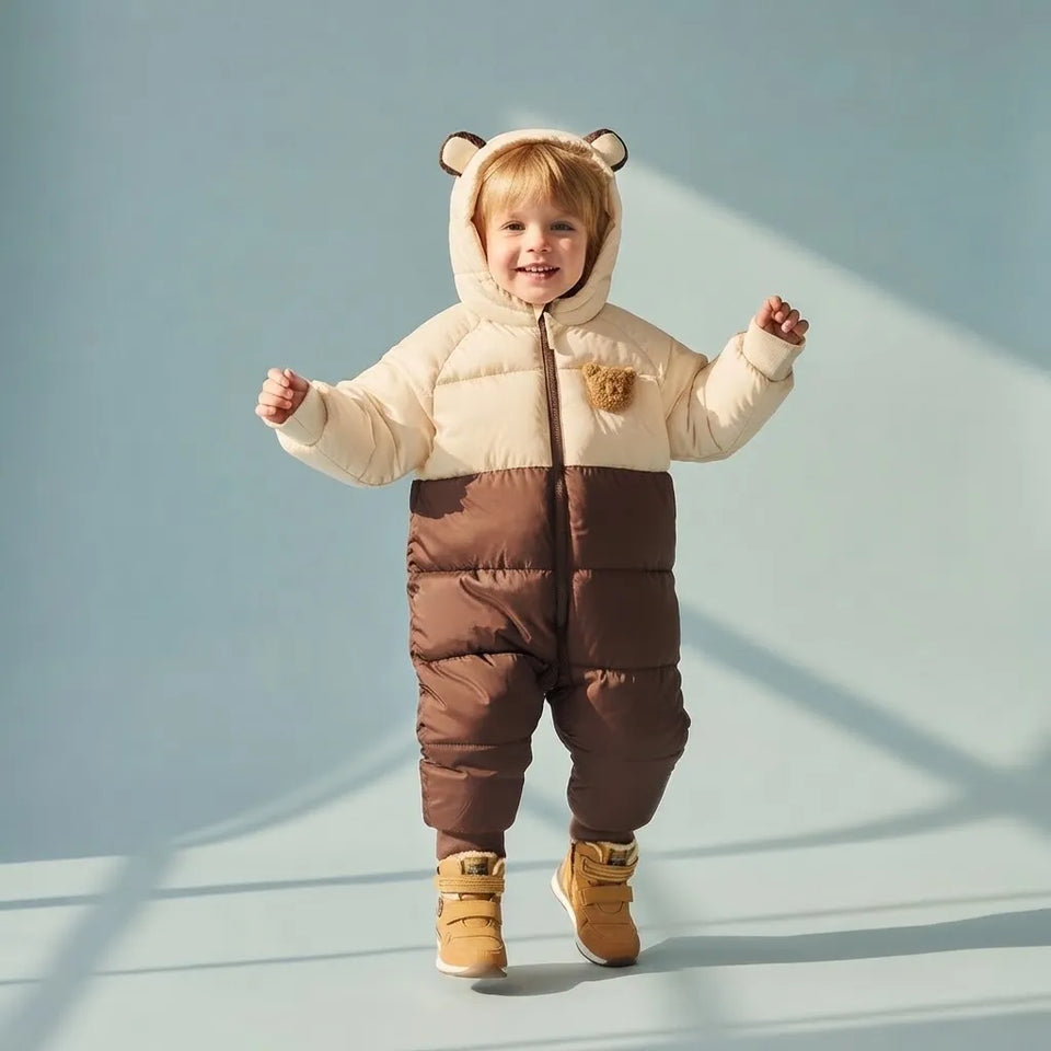 A toddler wearing a brown and cream bear-themed puffer snowsuit with a hood featuring bear ears and a small bear patch on the chest, standing against a light blue studio background.