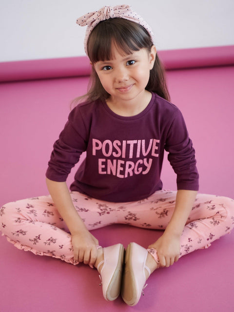 Young girl sitting on a pink surface wearing a 'Positive Energy' shirt.