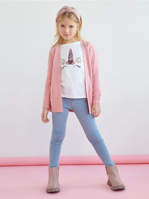 Young girl wearing a pink cardigan, white t-shirt with a unicorn design, blue jeans, and gray boots on a pink background.
