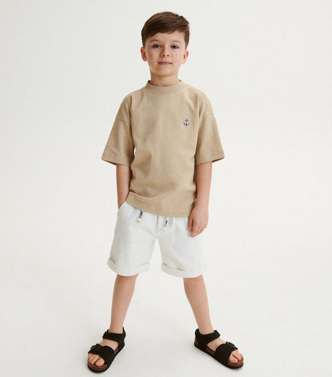 Young boy wearing a beige t-shirt, white shorts, and black sandals on a white background