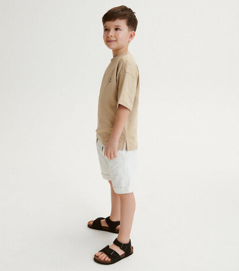 Young boy wearing a beige t-shirt, white shorts, and black sandals on a light gray background