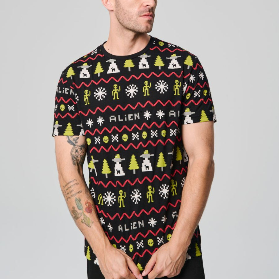 A man wearing a black short-sleeve t-shirt with a colorful 'Alien' themed holiday pattern, posed against a solid gray studio background.