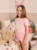 Young girl in pink pajamas with a daisy design, sitting on a bed with a teddy bear and butterfly decorations.