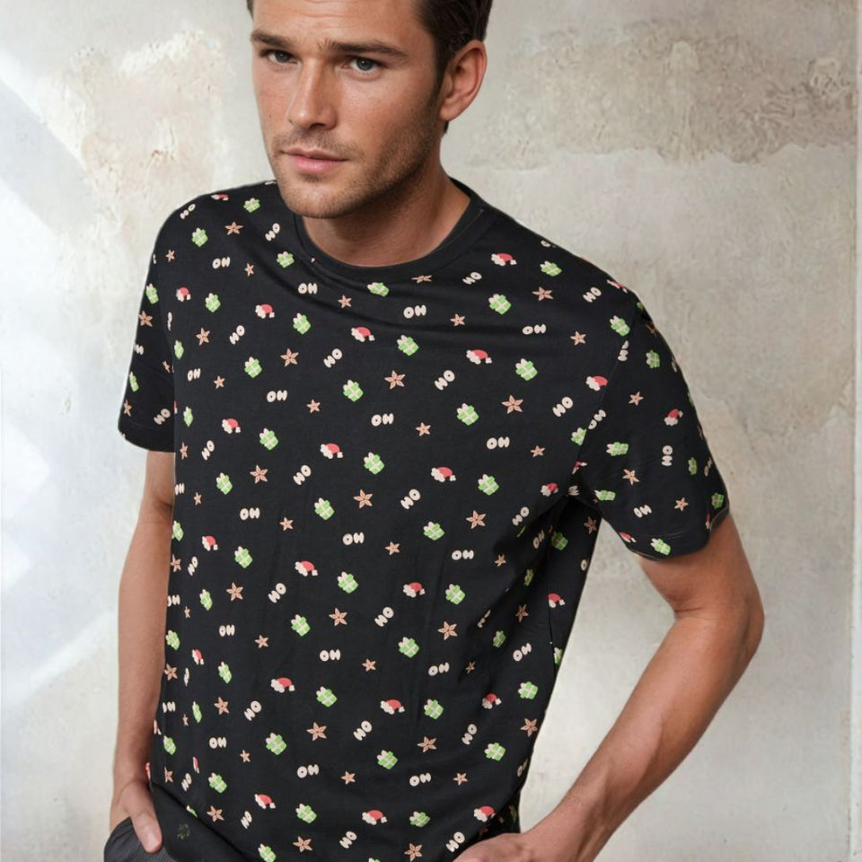 A man wearing a black short-sleeve crewneck t-shirt with a colorful retro video game pattern of green aliens, pink and white stars, and white score marks, posed against a textured concrete wall.