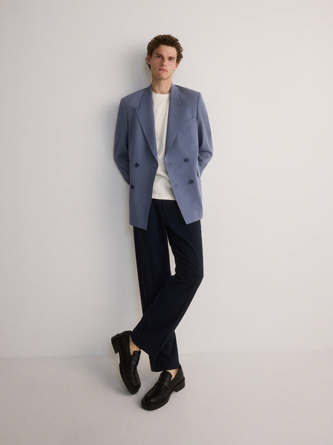 Man wearing a light blue blazer, white shirt, black pants, and black shoes on a plain background