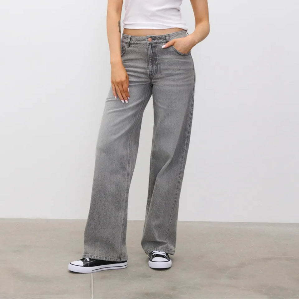 A person wearing gray wide-leg jeans, a white cropped top, and black-and-white sneakers, standing against a plain light gray wall and concrete floor.
