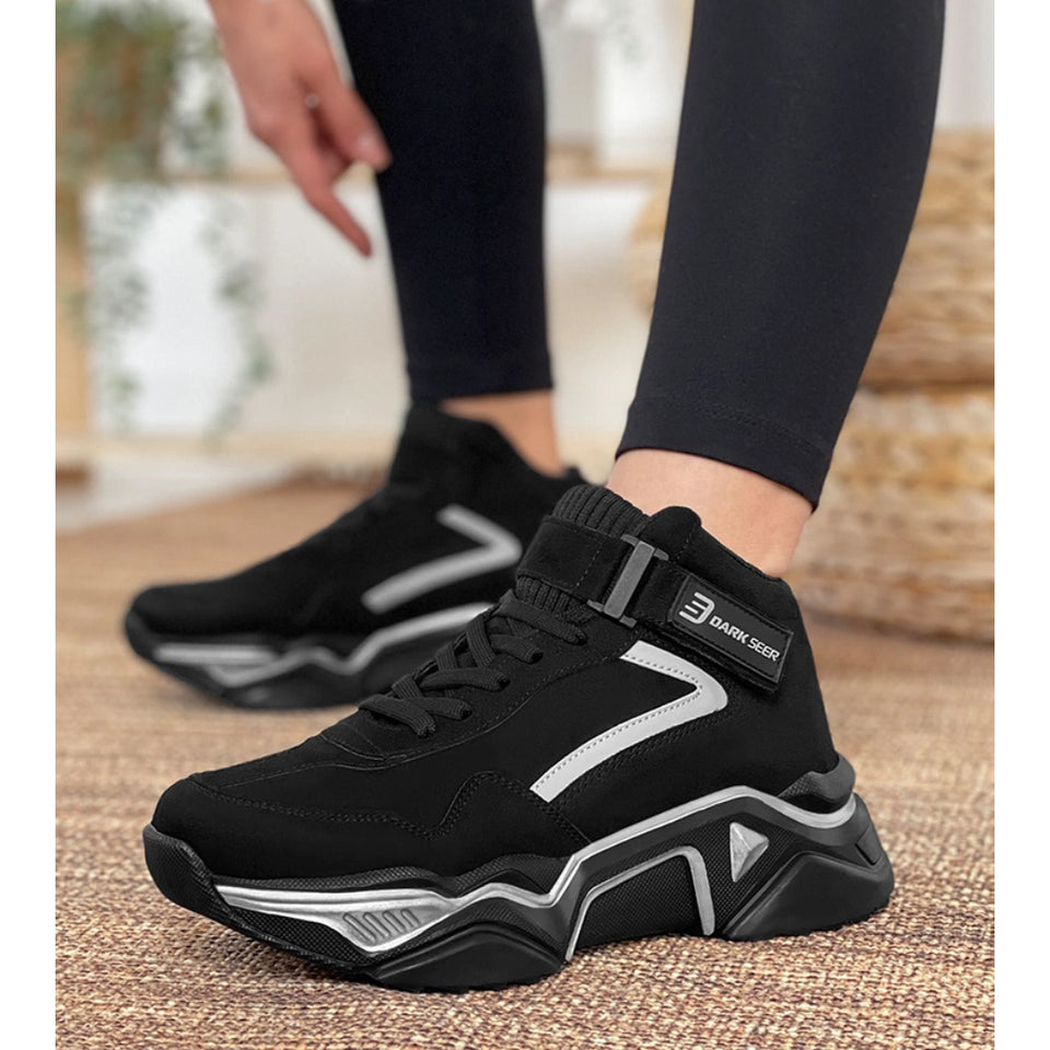 A person wearing black high-top sneakers with chunky silver and black geometric soles, standing on a textured beige floor. The shoes have black laces, a black ankle strap with a visible 'DAVID GEER' brand tag, and a black upper with silver graphic accents.