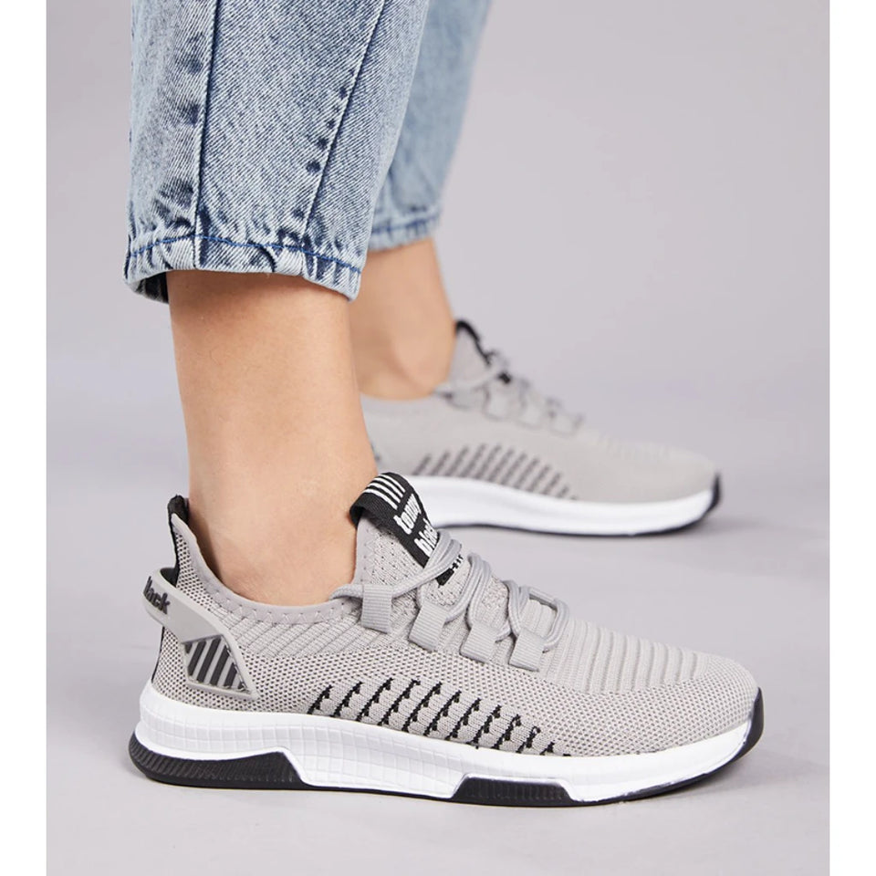 A pair of light gray knit athletic sneakers with black and white soles, shown on a model wearing light-wash blue jeans against a neutral gray studio background.