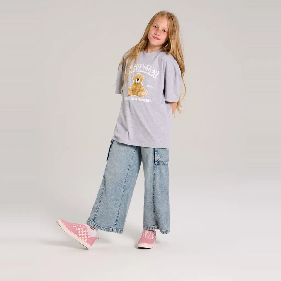 A young girl stands in a studio wearing a light gray graphic t-shirt with a teddy bear design, wide-leg light blue jeans, and pink sneakers.