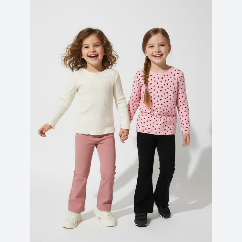 Two young girls smiling and holding hands, wearing matching long-sleeve tops and pants. The girl on the left wears an off-white knit top with dusty pink pants, and the girl on the right wears a pink top with black polka dots and black pants.