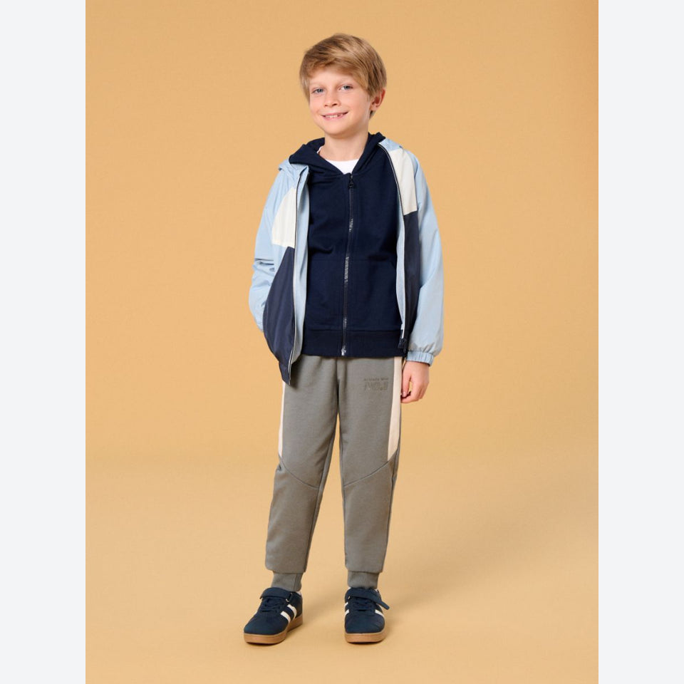 A young boy stands against a solid beige studio background wearing a color-blocked zip-up jacket in light blue, navy, and white, paired with gray color-blocked sweatpants and navy blue slip-on shoes.