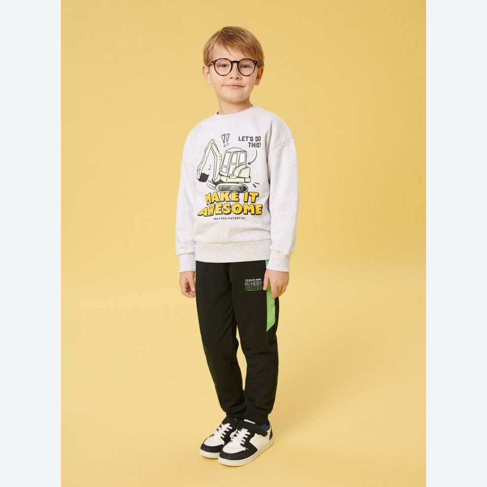 A young boy wearing a light gray graphic sweatshirt, black pants, and black-and-white sneakers, standing against a solid yellow background.