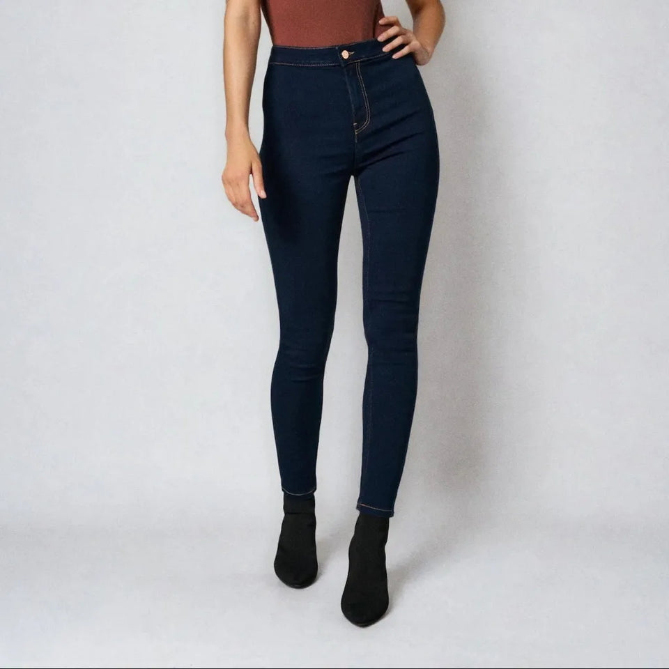 A model wearing dark navy blue high-waisted skinny jeans, paired with a rust-colored top and black ankle boots, standing against a light gray studio background.