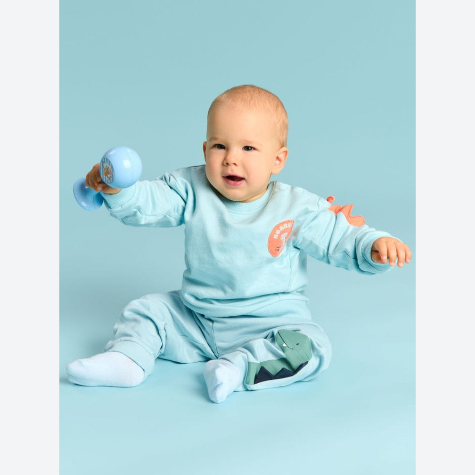 A baby sitting on a light blue studio background, wearing a matching light blue long-sleeve top and pants set with dinosaur-themed graphics, while holding a small blue toy rattle.