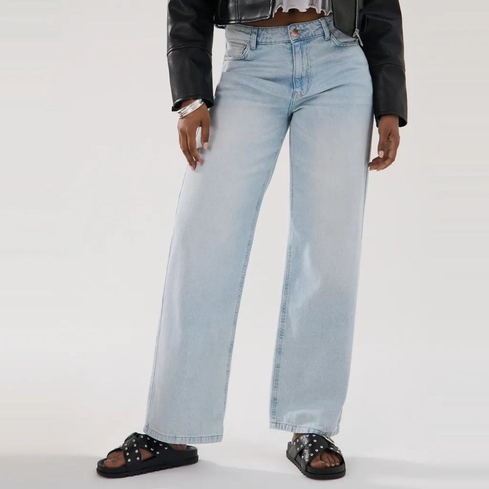 A person wearing light-wash wide-leg denim jeans, styled with a black leather jacket and black studded slide sandals, photographed against a clean white studio background.