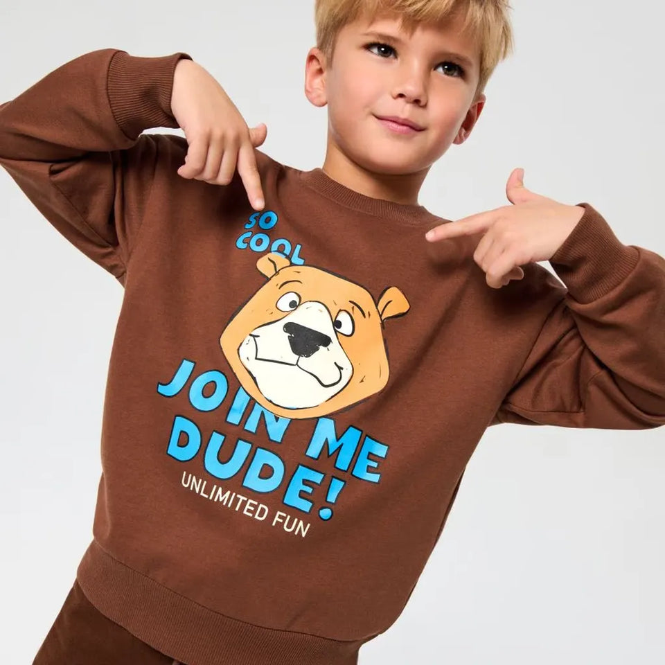 A young boy wearing a brown sweatshirt with a cartoon bear graphic and the text 'JOIN ME DUDE! UNLIMITED FUN' in blue and white, pointing at the design with both hands against a light gray background.